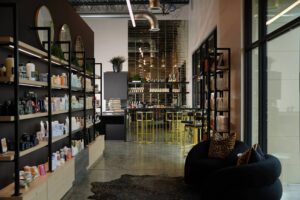 Modern Edwardsville salon interior with shelves of hair products, black lounge chairs, a rug, and a bar area featuring gold stools and decorative lights in the background. - Hollywood Hair Salon and Spa in Centralia, IL