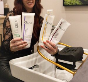 A person holds four CRLAB hair care product tubes near a salon sink and chair. - Hollywood Hair Salon and Spa in Centralia, IL