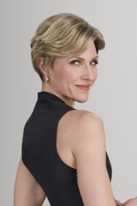 A woman with short blonde Hollywood Hair wearing a sleeveless black top looks over her shoulder and smiles against a plain light gray background. - Hollywood Hair Salon and Spa in Centralia, IL