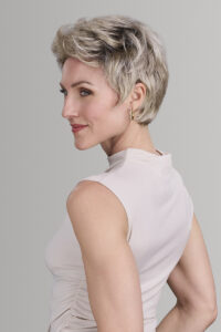 Woman with short, wavy blonde hair styled in Hollywood Hair by Jon Renau, wearing a sleeveless light beige top, looking over her shoulder against a plain gray background. - Hollywood Hair Salon and Spa in Centralia, IL