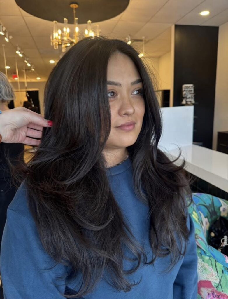 A woman with long, voluminous dark hair styled in loose layers sits in a modern salon, wearing a blue sweatshirt. A hand adjusts her hair. - Hollywood Hair Salon and Spa in Centralia, IL