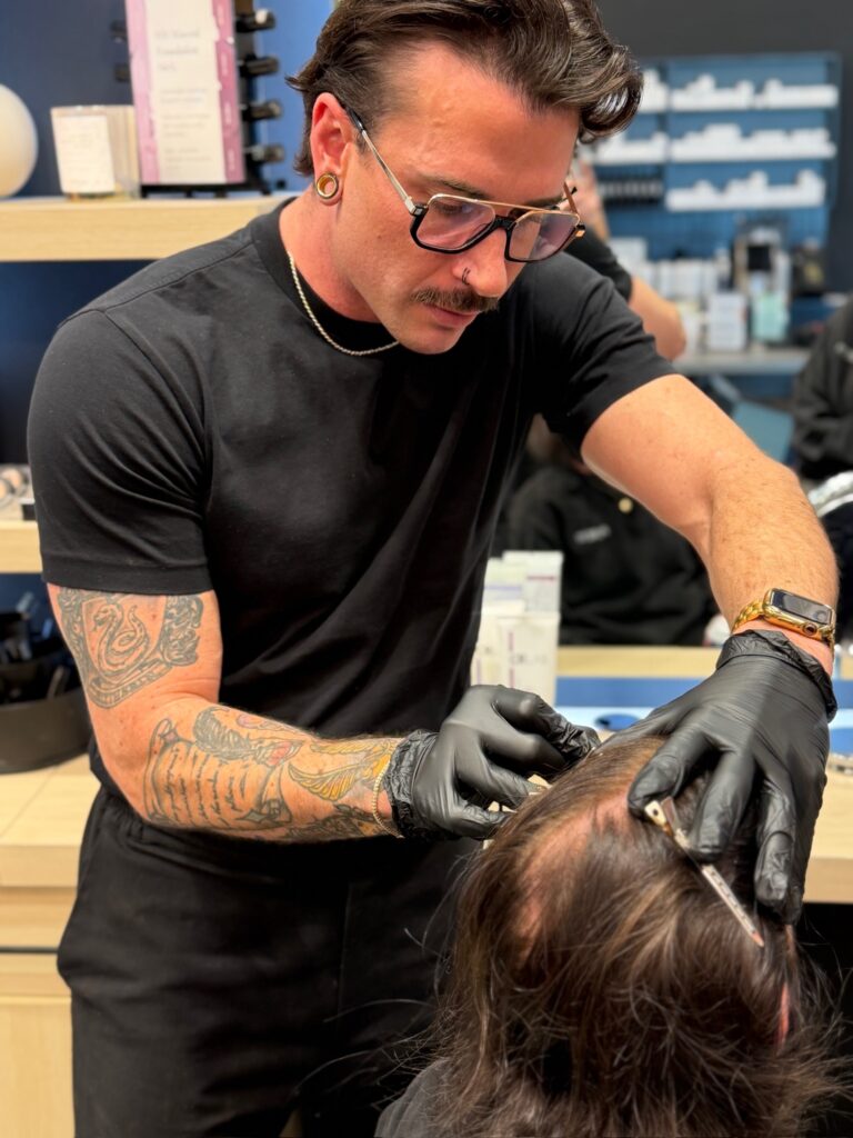 A tattooed man in black clothing and gloves administers an injection to a person’s scalp in a clinical setting. - Hollywood Hair Salon and Spa in Centralia, IL