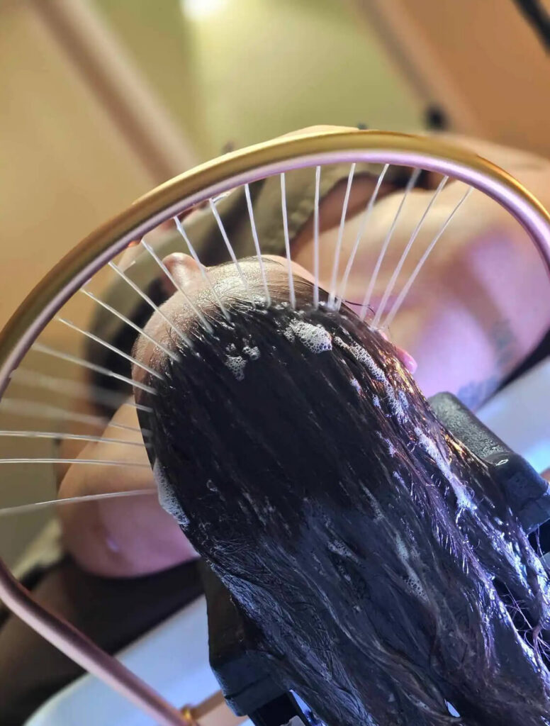 Person having their hair washed under a ring-shaped showerhead with water streams and soap suds visible on their wet hair. - Hollywood Hair Salon and Spa in Centralia, IL