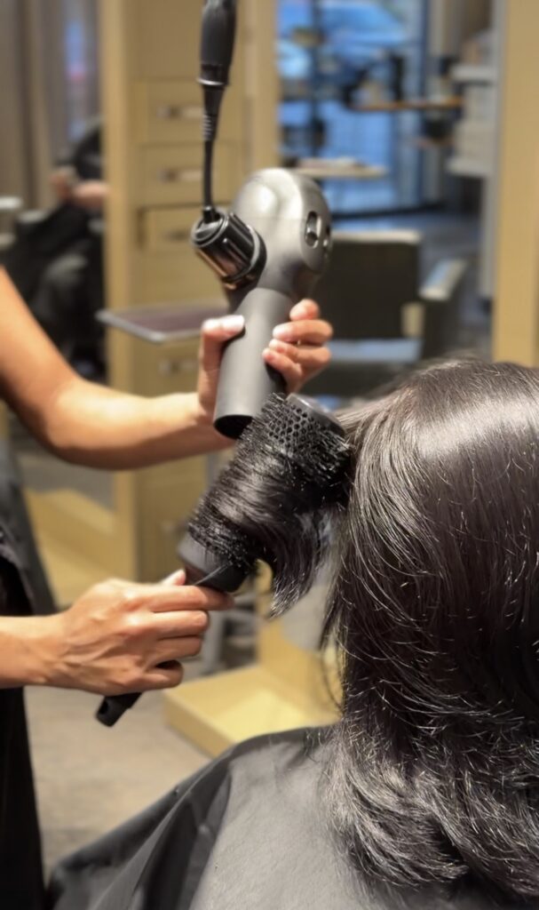A person blow-dries another person's shoulder-length dark hair using a round brush and hair dryer in a salon setting. - Hollywood Hair Salon and Spa in Centralia, IL
