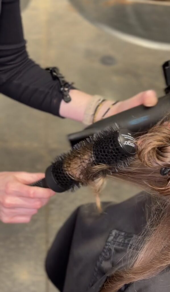 A person uses a round brush and blow dryer to style someone's light brown hair in a salon. - Hollywood Hair Salon and Spa in Centralia, IL