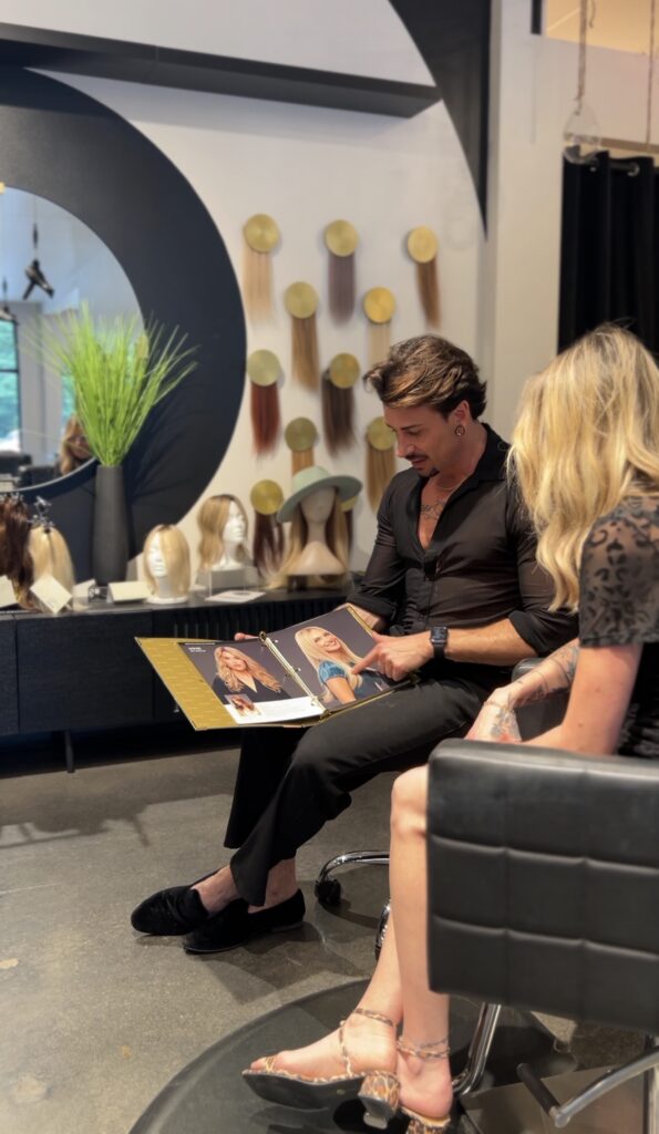 A man and woman sit in a salon, looking at a booklet with hair color options. Wigs and hairpieces are displayed on the shelves behind them. - Hollywood Hair Salon and Spa in Centralia, IL
