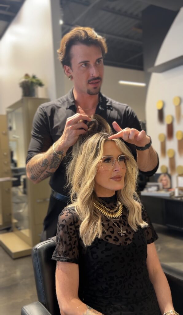 A hairstylist works on a woman's hair in a modern salon; the woman is seated, wearing glasses and a black outfit, while the stylist stands behind her. - Hollywood Hair Salon and Spa in Centralia, IL