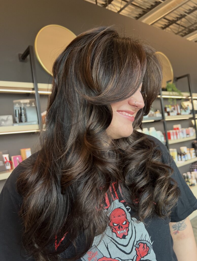 A person with long, layered, wavy brown hair smiles in a salon, wearing a black graphic t-shirt. Salon products and shelves are visible in the background. - Hollywood Hair Salon and Spa in Centralia, IL