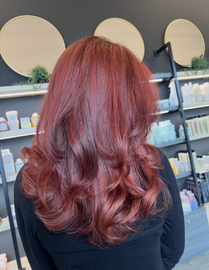 Person with shoulder-length, wavy, vibrant red hair facing away, standing in a modern salon with shelves of hair products and round mirrors on the wall. - Hollywood Hair Salon and Spa in Centralia, IL