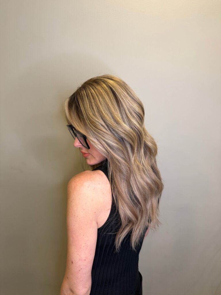 A woman with long, wavy blonde hair wearing black glasses and a sleeveless black top stands against a plain beige wall, facing away from the camera. - Hollywood Hair Salon and Spa in Centralia, IL