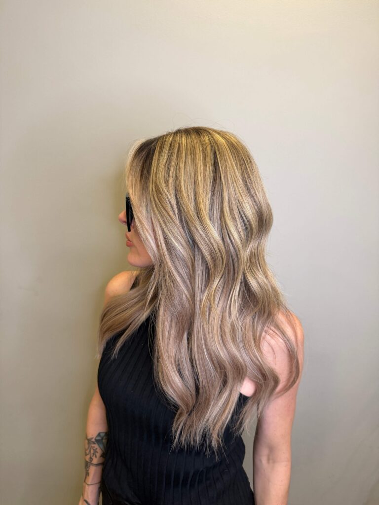 A woman with long, wavy, blonde hair wearing a sleeveless black top looks to the side against a plain beige background. - Hollywood Hair Salon and Spa in Centralia, IL