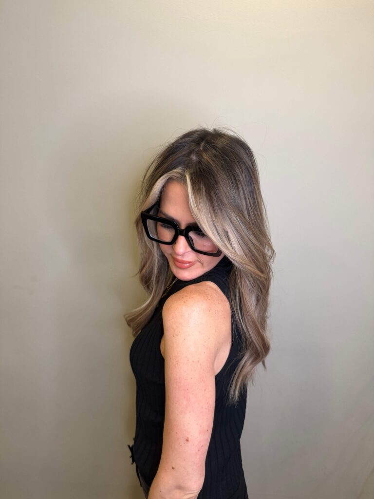 Woman with long, wavy blonde hair wearing black glasses and a sleeveless black top stands against a plain beige wall, looking down and to the side. - Hollywood Hair Salon and Spa in Centralia, IL