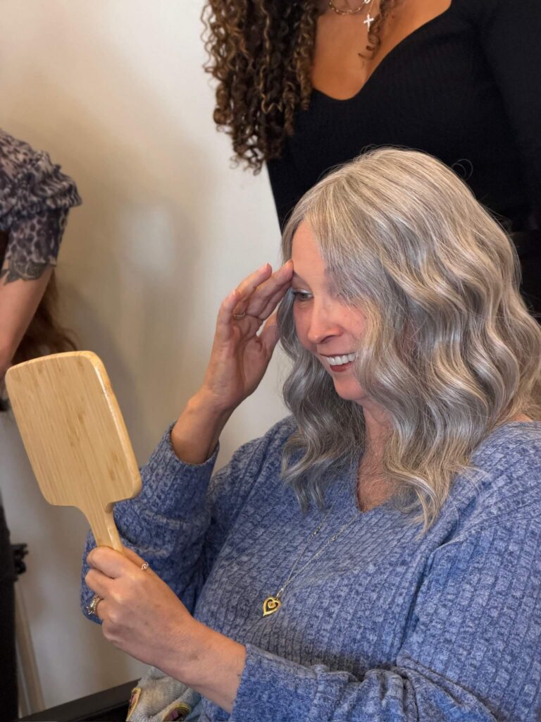 A woman with long gray hair smiles while looking at herself in a handheld mirror. Two people stand nearby, partly visible. - Hollywood Hair Salon and Spa in Centralia, IL