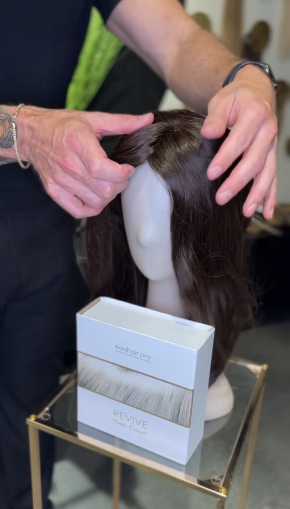 A person adjusts a brown wig on a mannequin head next to a box labeled "easihair pro REVIVE" on a small glass table. - Hollywood Hair Salon and Spa in Centralia, IL